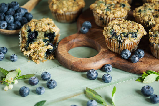 Fresh blueberry muffins with oat streusel topping and raw blueberries spilling from a wooden spoon. Selective focus on muffins on cutting board with blurred background.
