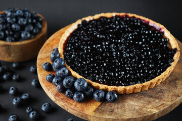 Opened blueberry tart on a wooden stand next to fresh blueberries on a dark background