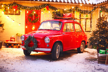 Red retro car in snow-covered courtyard of house decorated with pine branches and xmas lights