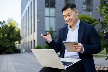 Smiling young Asian male businessman in a suit sitting outside an office building, holding a laptop...