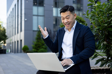 A happy young Asian male businessman is sitting outside an office building and smilingly looking at...
