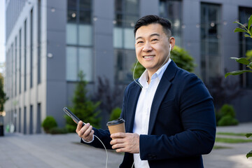 Portrait of an Asian young male office worker wearing headphones and holding a phone and a cup of coffee resting sitting outside an office center and smiling at the camera