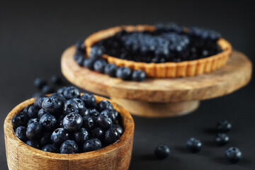 Fresh blueberries in a wooden bowl next to blueberry tart on a wooden stand on a dark background