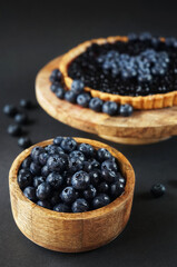 Fresh blueberries in a wooden bowl next to blueberry tart on a wooden stand on a dark background