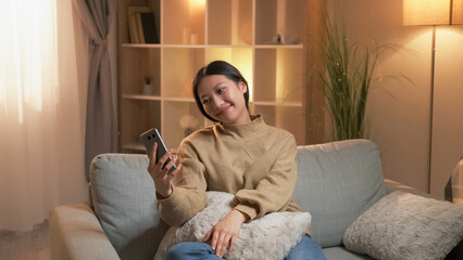 Virtual call. Happy woman. Mobile communication. Relaxed smiling female looking smartphone sitting sofa in living room interior. - 787206275