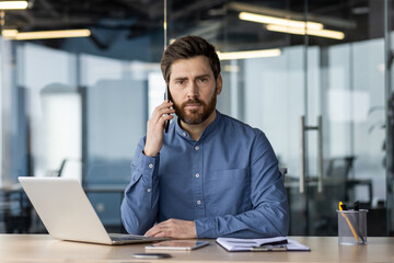 Portrait of a serious young businessman sitting in the office at the table in front of the camera and talking on the phone