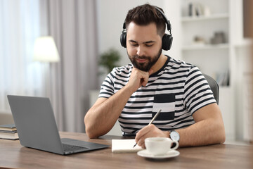 Young man in headphones writing down notes during webinar at table in room