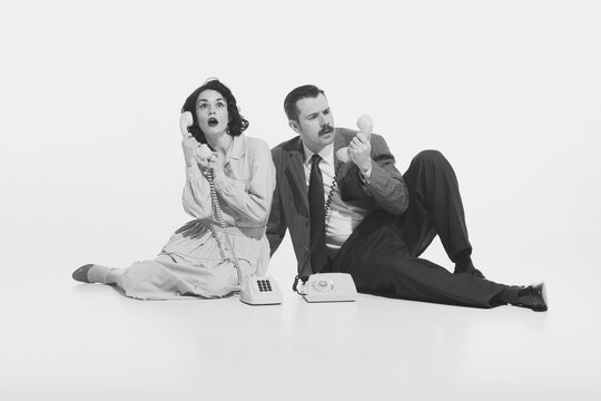Monochrome i,age of elegant young woman and stylish man sitting on floor and expressively talking on vintage rotary phone. Concept of retro and vintage, fashion, business, human emotions, style