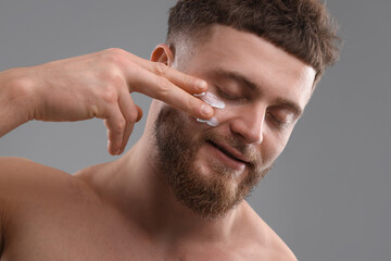 Obraz premium Handsome man applying moisturizing cream onto his face on grey background
