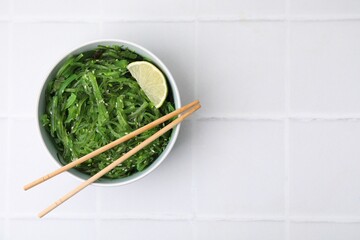 Tasty seaweed salad in bowl served on white tiled table, top view. Space for text