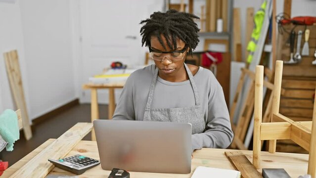 Focused young woman with dreadlocks using laptop at her woodworking workshop, depicting creativity and entrepreneurship.