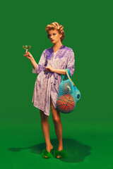 Young woman in silky bathrobe with hair curlers holding glass of cocktail sport balls against vibrant green background. Concept of sport, competition, active lifestyle, hobby, recreation. Ad