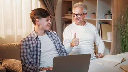 Good job. Supportive father. Family education. Cheerful dad showing like gesture to son with great homework on laptop home interior.