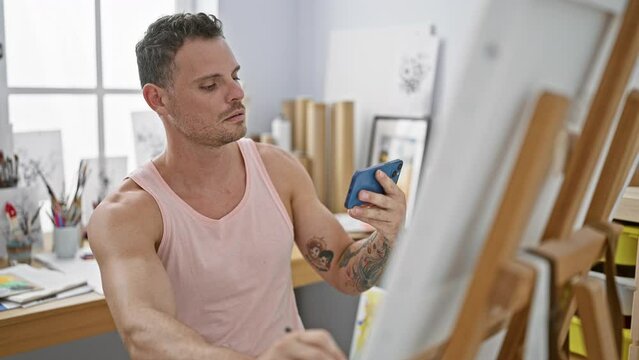 A young adult man with tattoos carefully examines his phone in a bright art studio.