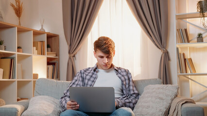 Online work. Male worker. Digital technology. Serious man browsing laptop sitting sofa in light home interior.