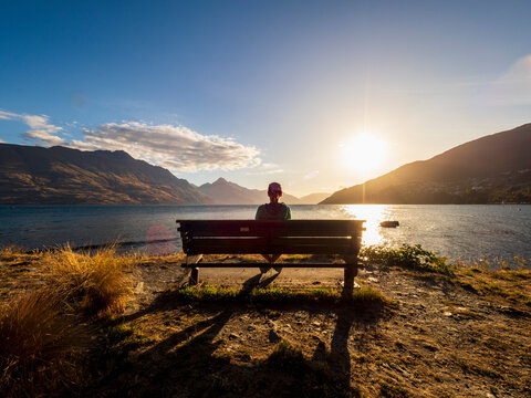 Rear view of woman sitting on bench, facing Lake Wakatipu at sunset