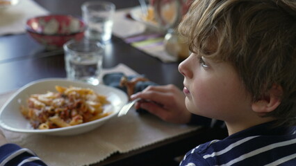 Pensive profile face of a small boy seated at lunch table with contemplative gaze. Child holding...