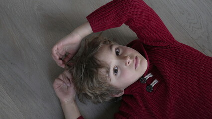 Pensive young boy plays with hair while looking at camera laid on floor looking from bellow. Child...