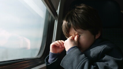 Tired young boy rubbing face and eyes while traveling by train. drowsy traveller child seated by...