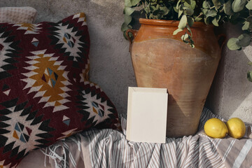 Blank greeting card, invitation mockup, craft envelope. Summer vacation still life, relaxation concept. Cozy bench with Mexican pattern cushions. Vase, eucalyptus branches, fresh lemons. Striped plaid