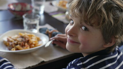 Young boy turning head toward camera seated at lunch table smiling. Child eating pasta food, turns head back to plate. Handsome portrait