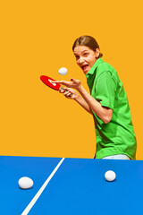 Funny woman in green shirt playing ping pong, excited expression against yellow studio background....