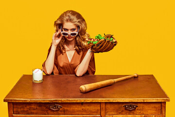 Stylish woman with sunglasses holds hotdog in baseball glove, with bat at wooden desk, against yellow studio background. Concept of sport, active lifestyle and healthy eating, contemporary art.