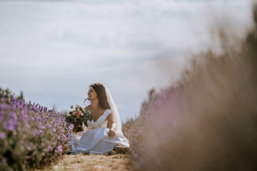 Valmiera, Latvia- July 28, 2024 - A bride sits amidst lavender, gazing away thoughtfully, veiled in soft light with blooms and sky in the backdrop.