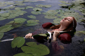 Red haired woman in red medieval dress holding long sword floating in lake with lily pads. Fantasy...