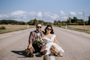 Valmiera, Latvia- July 28, 2024 - A bride and groom sit in the middle of a deserted road, the bride holding a bouquet, both wearing sunglasses and smiling.