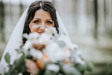 Valmiera, Latvia- July 28, 2024 - Bride peeks over her bouquet, veil draped, with a joyful gaze in...