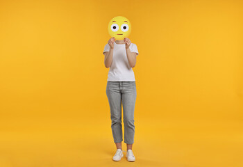 Woman covering face with surprised emoticon on yellow background