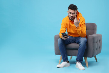 Emotional young man using smartphone on armchair against light blue background, space for text