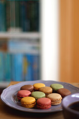 Obraz na płótnie Canvas Purple plate filled with pastel macarons, cup of tea or coffee, vintage books and reading glasses on the table. Colorful bookcase in the background. Selective focus.