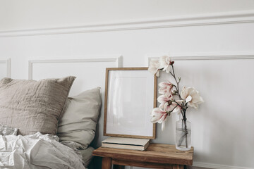 Empty vertical wooden picture frame mockup, old books. Wooden night stand with fluted glass vase. Blooming magnolia tree branches. Scandinavian interior, bedroom. White wall background, side view