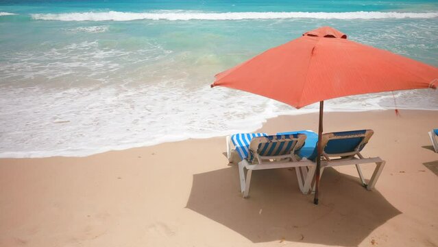 Two empty blue sun loungers under the red parasol on the sandy beach near the sea water with big waves. Rest and relaxation summer holiday scene