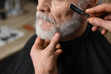 Professional barber shaving client's beard with blade in barbershop, closeup