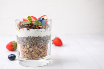 Tasty granola with berries, yogurt and chia seeds in glass on white tiled table, space for text
