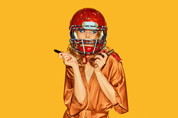 Beautiful woman in elegant attire wearing football helmet makes makeup against yellow studio background. Concept of woman in football, sport and fashion, style and beauty. Ad