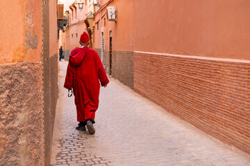 man in traditional long attire walks down street along high wall Red, City Marrakech, back view,...