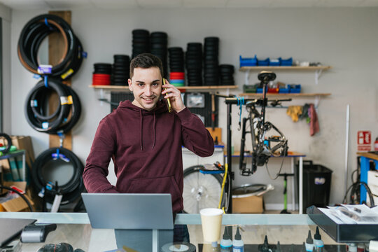 The bike mechanic is answering the phone with a smile, looking at the camera with a laptop in front of him on the counter of his bike workshop.