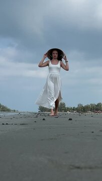 4K Young woman on an empty beach in a white dress walks along the sand holding her shoes. European girl wearing Vietnamese hat