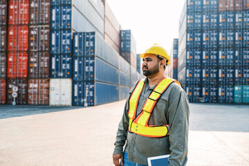 An attractive Indian male industrial engineer in yellow hard hat, safety vest with a blurred container yard in background. Working in the logistics center. Inspector, supervisor in container terminal.