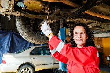 Portrait of a Caucasian female mechanic in a red uniform standing under the car bottom for inspecting in the garage. A woman smiling while holding a wrench. Car repair service. Vehicle maintenance
