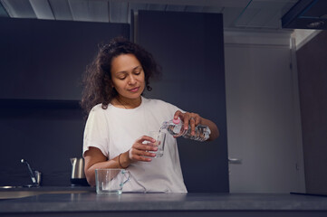 Young woman in pajamas, pouring clean fresh mineral water from bottle into a glass, drinking water...