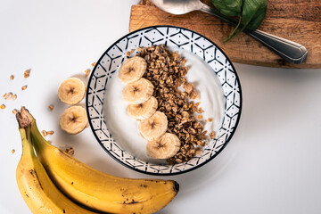 Breakfast bowl with yogurt, cereal and sliced pieces of banana with a wooden board with a spoon and two bananas around it
