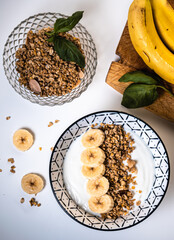 Overhead view of a breakfast bowl with yogurt, cereal and sliced ​​banana pieces with a wooden board two bananas and another glass bowl with cereals around
