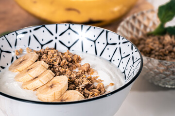 Breakfast bowl with yogurt, cereal and cut pieces of banana with bananas and another bowl with cereal out of focus in the background