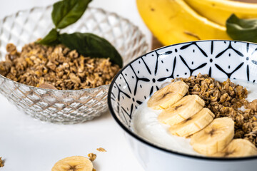 Breakfast bowl with yogurt, cereal and sliced ​​banana pieces with a glass bowl with cereal and bananas out of focus in the background
