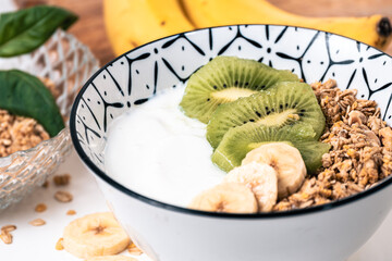 Breakfast bowl with yogurt, cereal and sliced pieces of kiwi and banana with bananas and another bowl with cereal out of focus in the background
